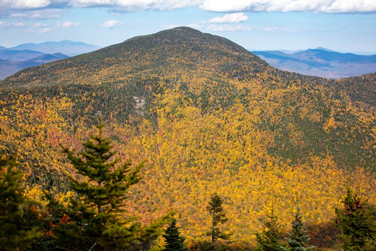 Mount Passaconaway covered in fall colors with forest and distant peaks under blue sky